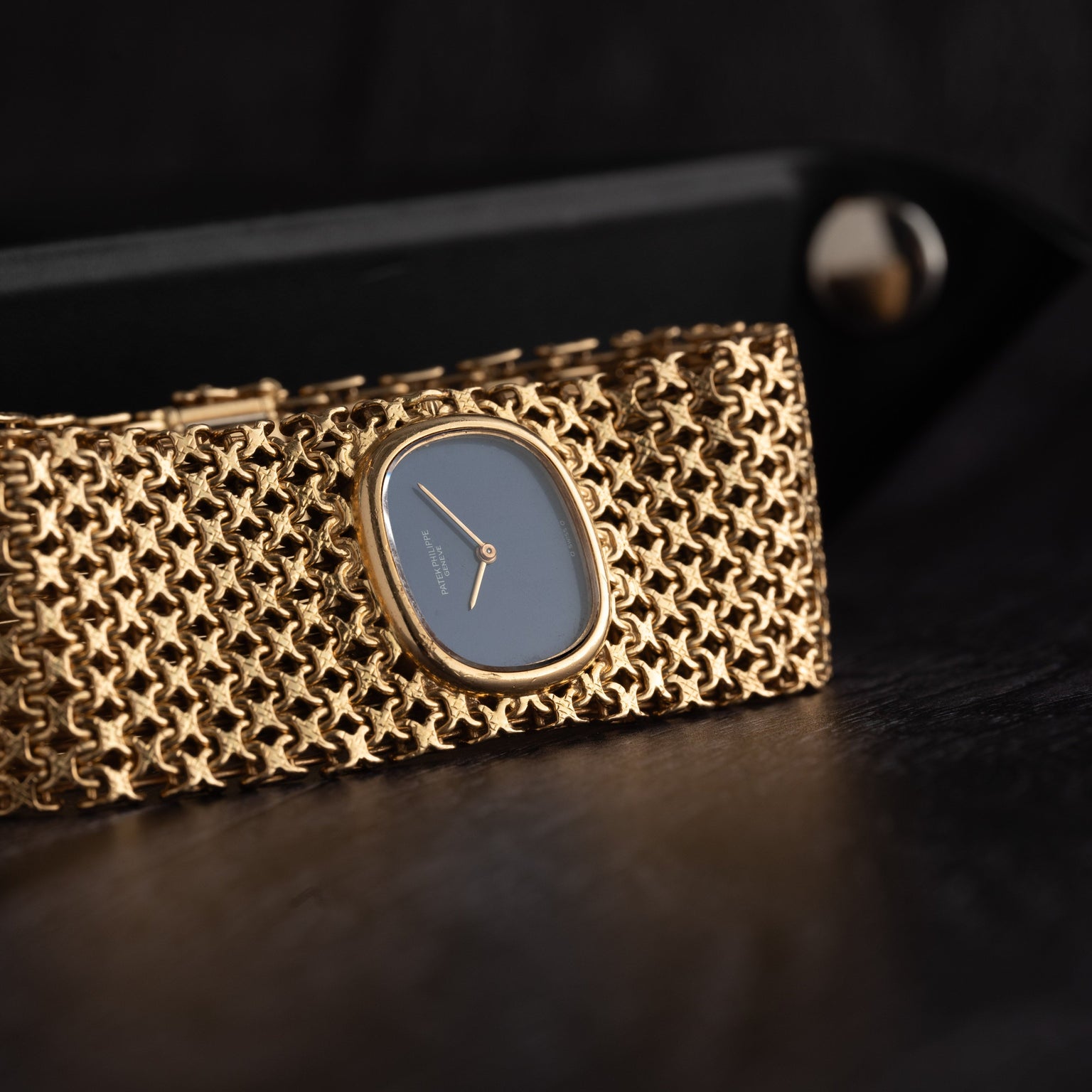 A close-up of the Patek Philippe Ellipse Ladies Woven Cuff watch with a blue face and gold mesh cuff, displayed on a dark surface.