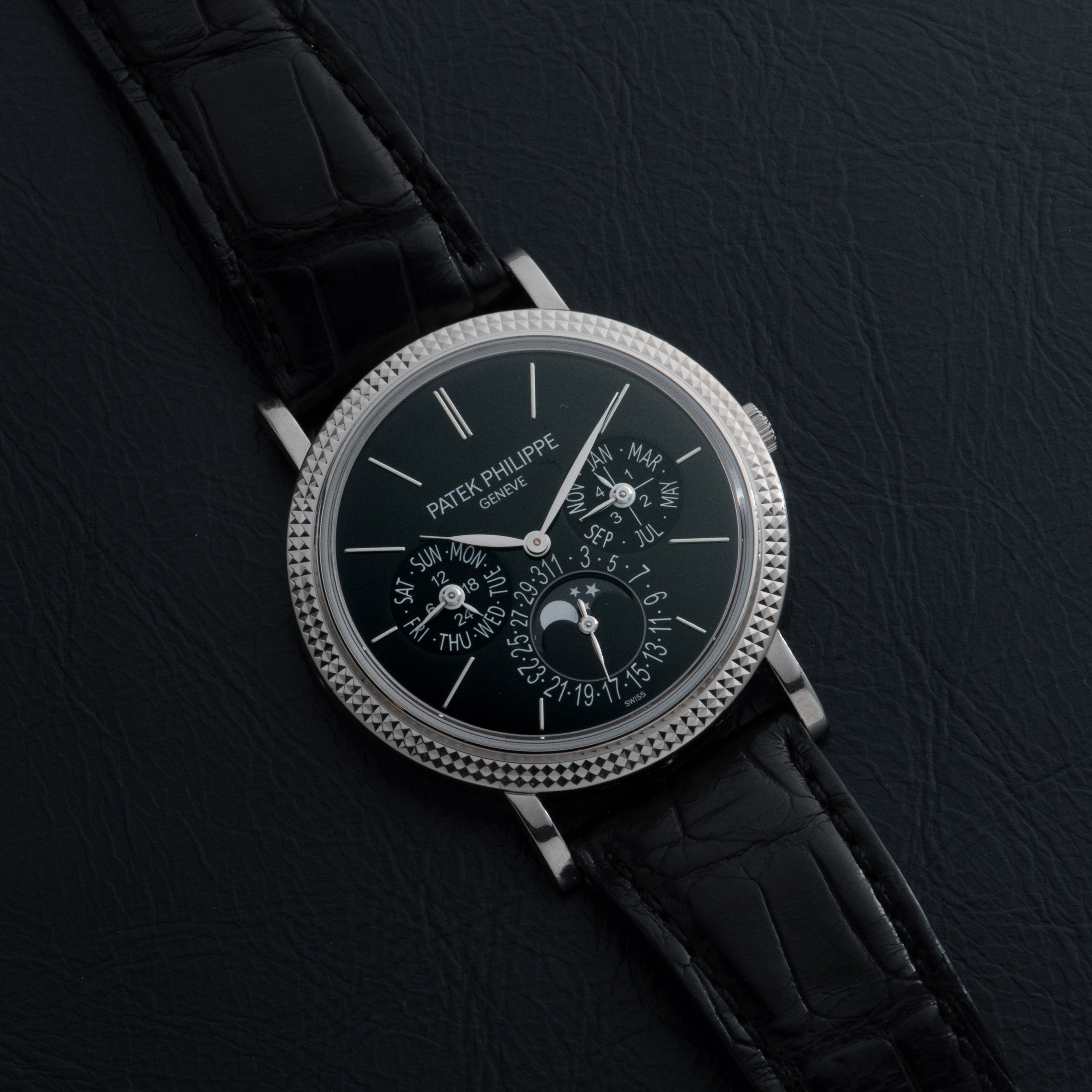 A luxurious black Patek Philippe Perpetual Calendar Moonphase wristwatch with a detailed dial and black leather strap displayed on a dark surface.