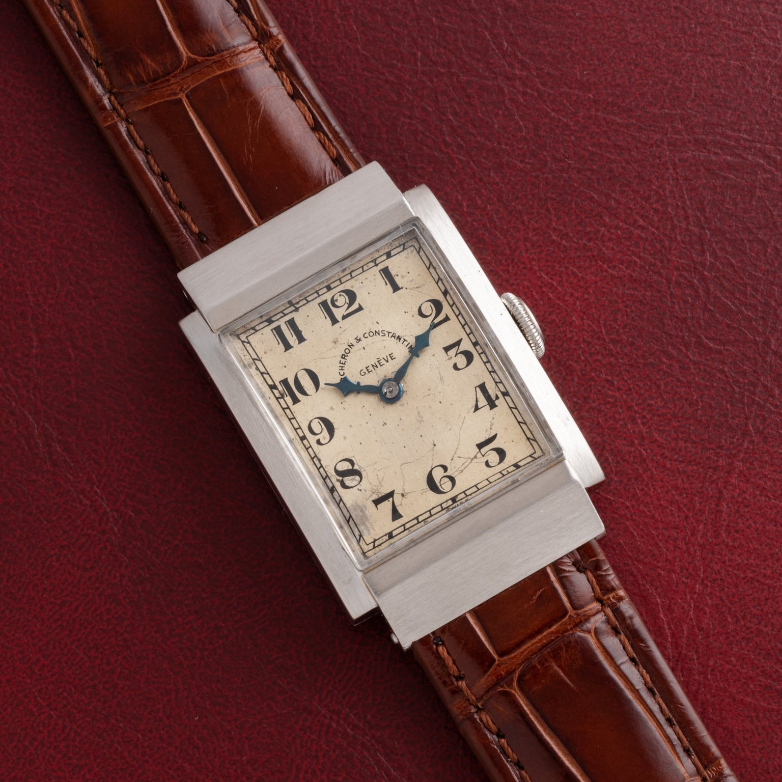 A Vacheron Constantin Rectangular Steel Dress Watch with a brown leather strap displayed on a textured burgundy surface.