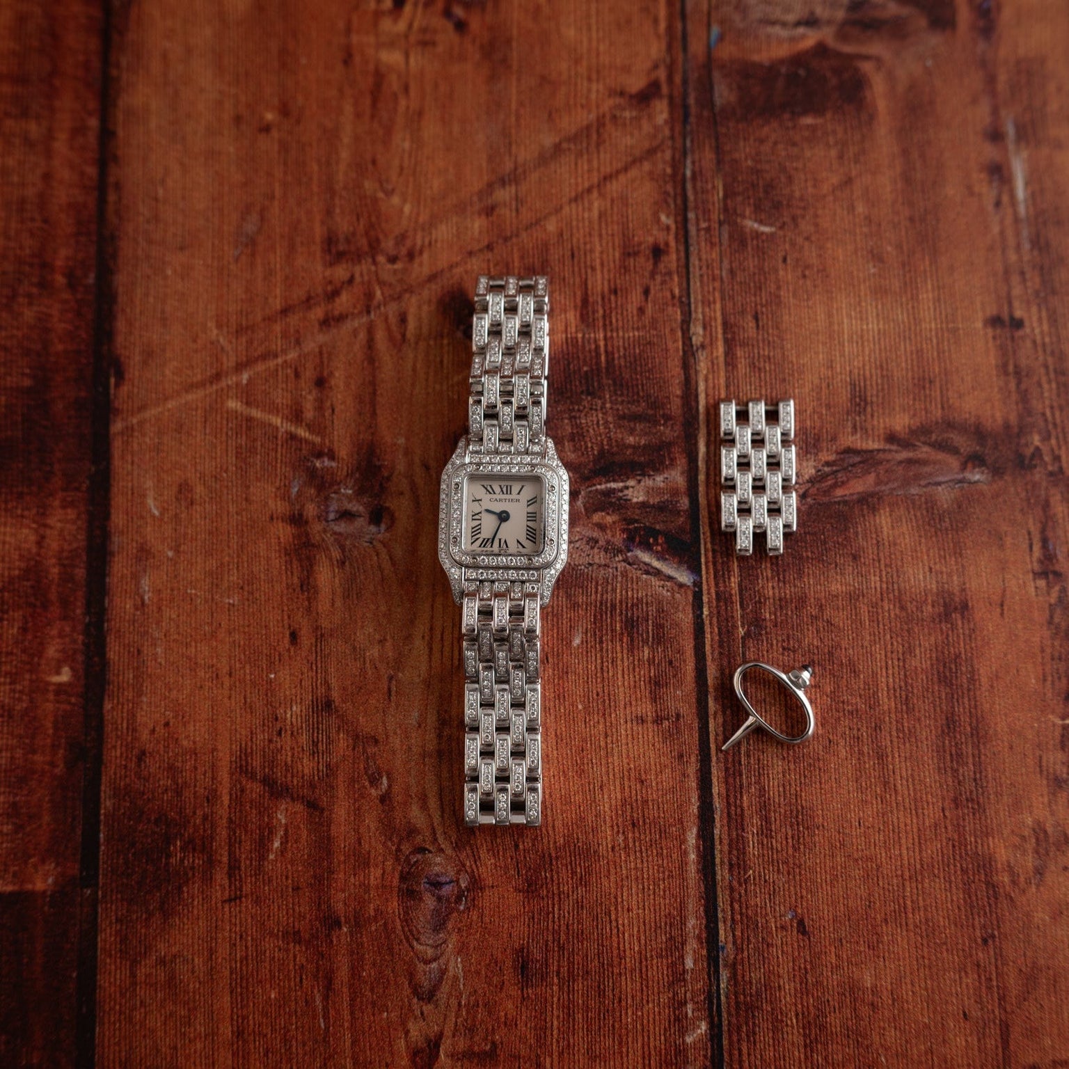 A Cartier Panthère Diamond silver wristwatch, extra watch links, and a small hoop earring rest on a wooden surface.