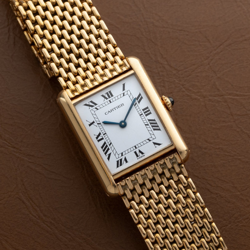 A Cartier Tank Louis watch with a gold case, white rectangular dial, and Roman numerals, displayed on brown leather.