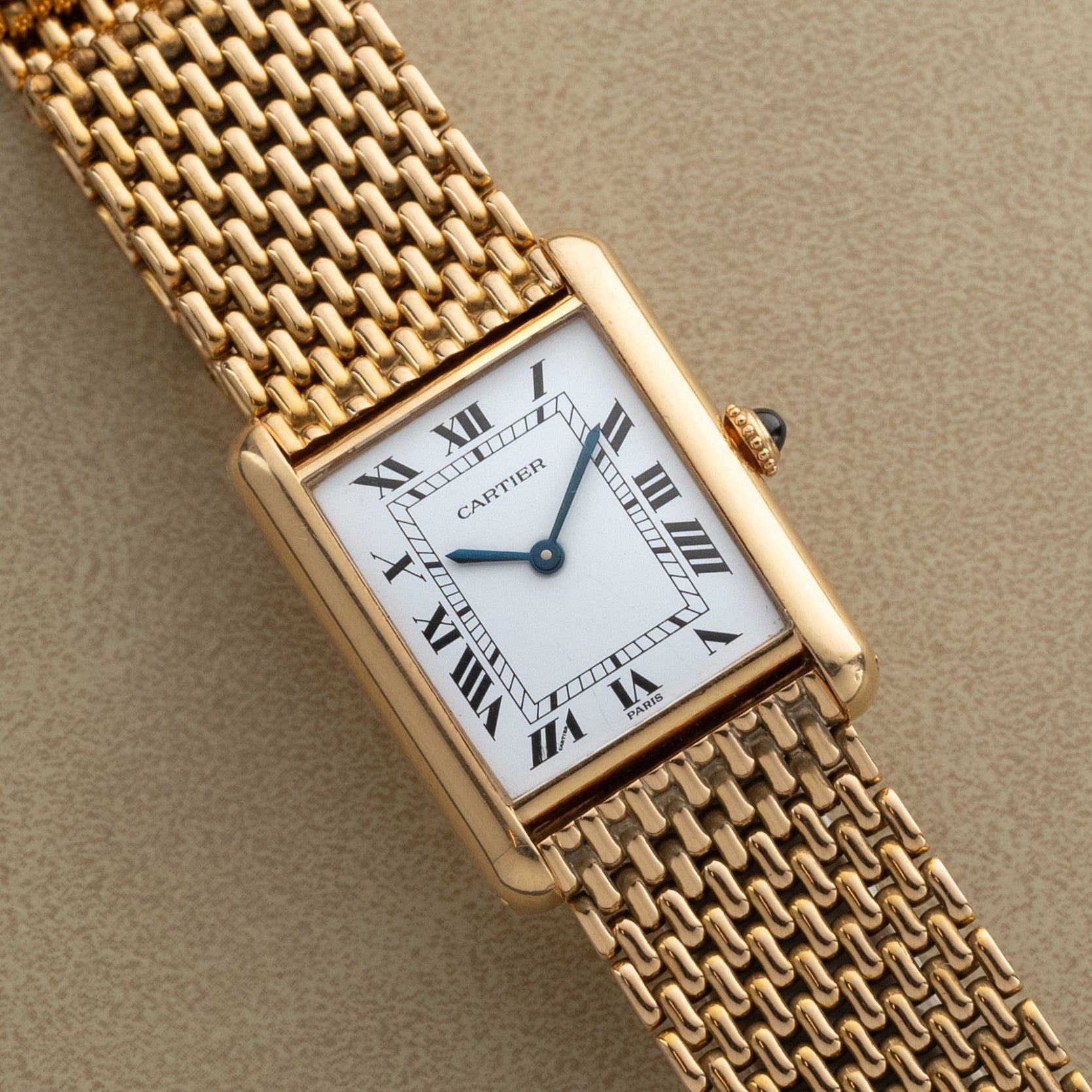 A Cartier Tank Louis gold wristwatch with a rectangular face, Roman numerals, and mesh bracelet rests on a beige surface.