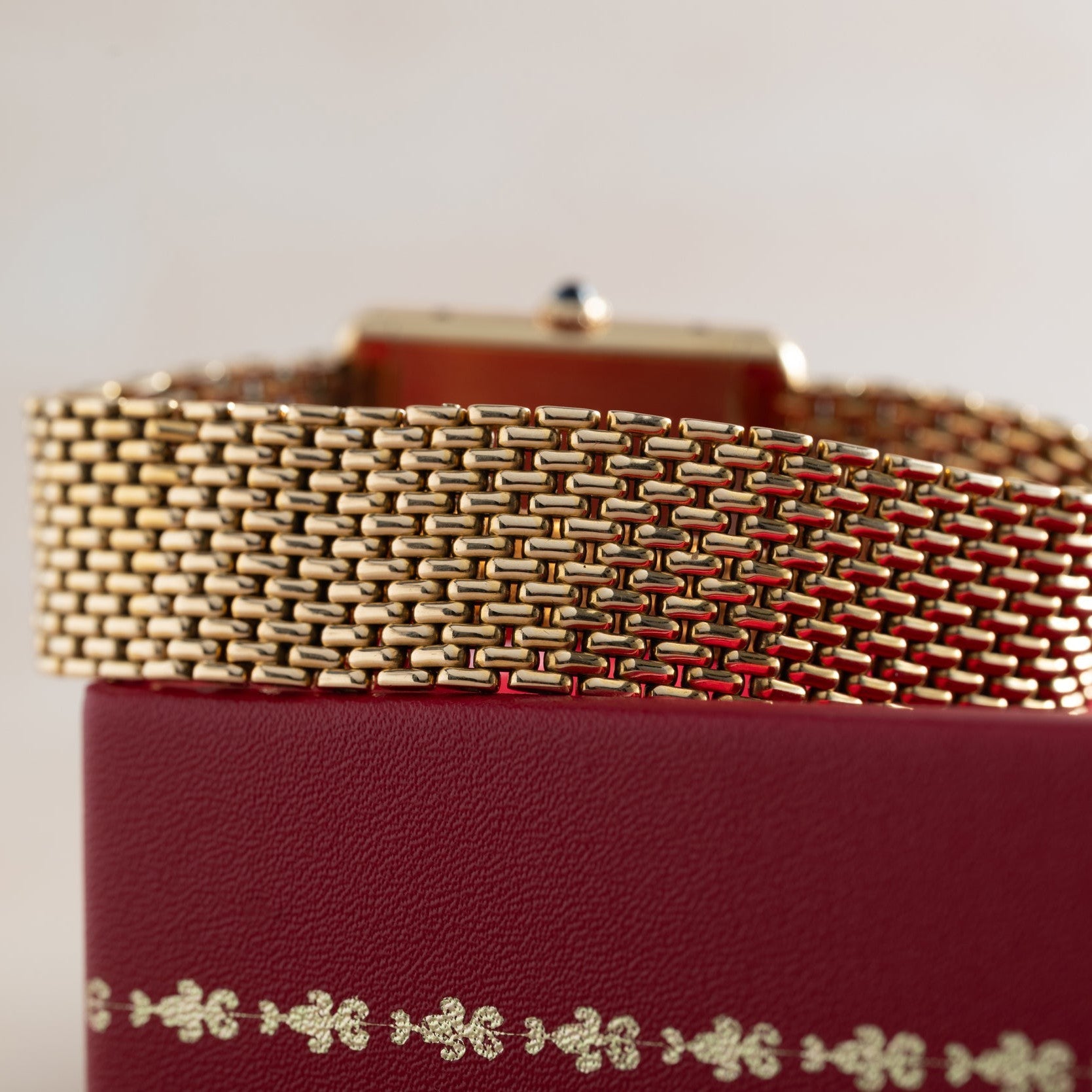 Close-up of the Cartier Tank Louis gold mesh watch band resting on a red textured box with gold decorative accents.