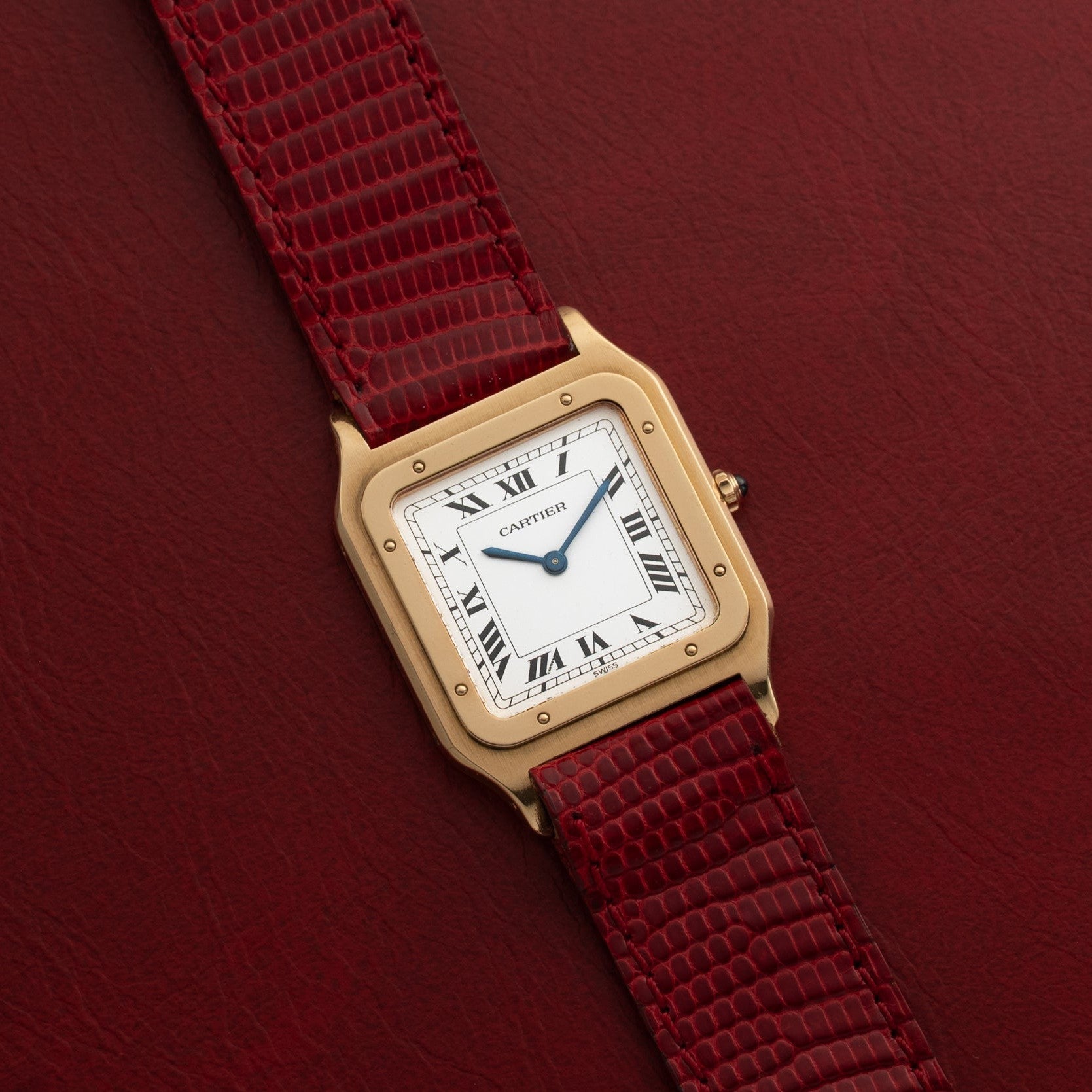 The Cartier Santos Dumont watch features a white Roman numeral dial and a red textured leather strap, displayed here on a red background.