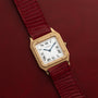 The Cartier Santos Dumont watch features a white Roman numeral dial and a red textured leather strap, displayed here on a red background.