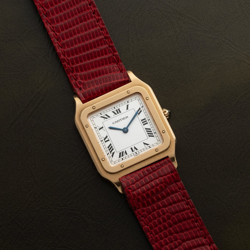 The Cartier Santos Dumont gold watch with a red leather strap is displayed on a dark, textured surface.