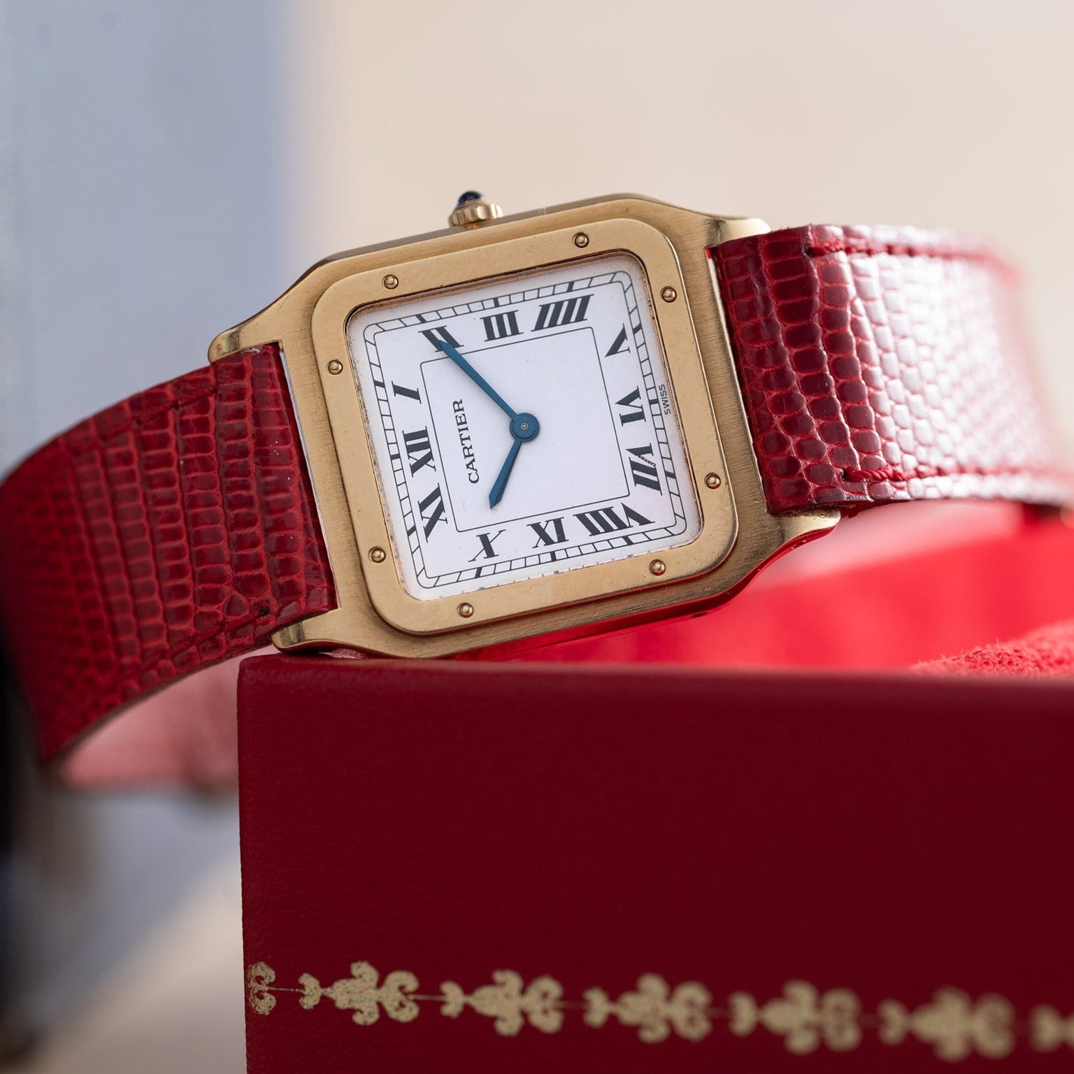 Cartier Santos Dumont gold watch featuring Roman numerals and a red leather strap, displayed on a red box.