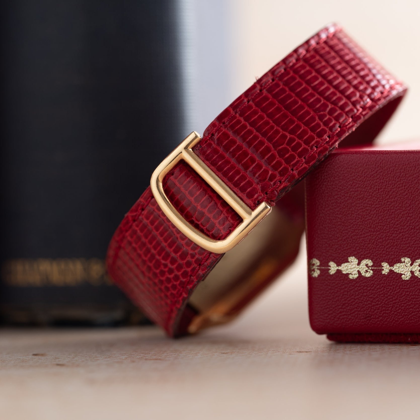 Close-up of a red textured Santos Dumont watch strap by Cartier, featuring a gold buckle, placed next to a red box and a dark book.