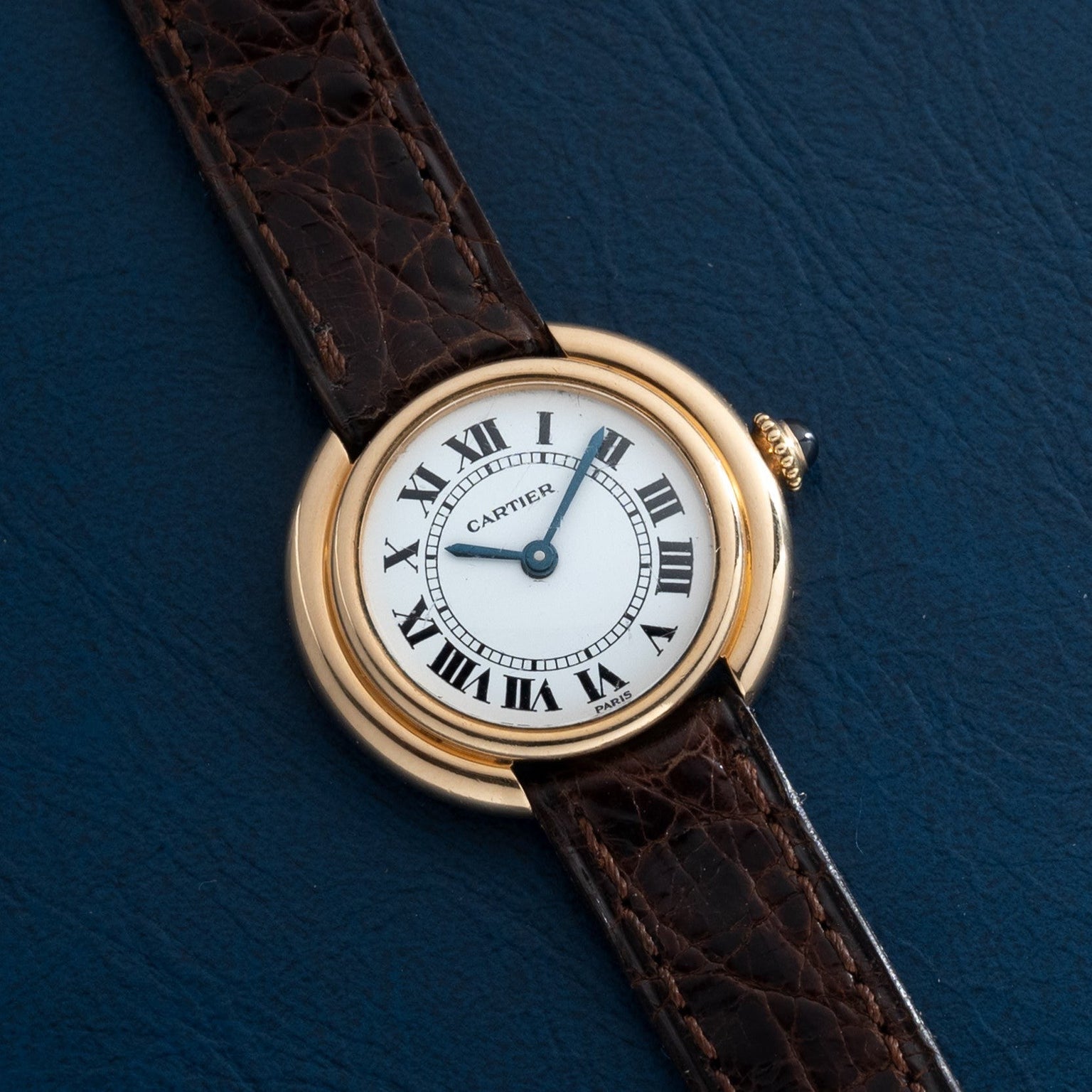A Cartier Vendome watch with a brown leather strap and Roman numeral dial, displayed on a dark blue surface.