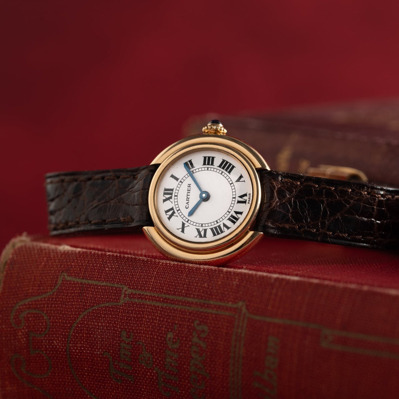 A Cartier Vendome gold watch with a black leather strap sits atop a red vintage book, set against a red background.