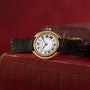 A Cartier Vendome gold watch with a black leather strap sits atop a red vintage book, set against a red background.