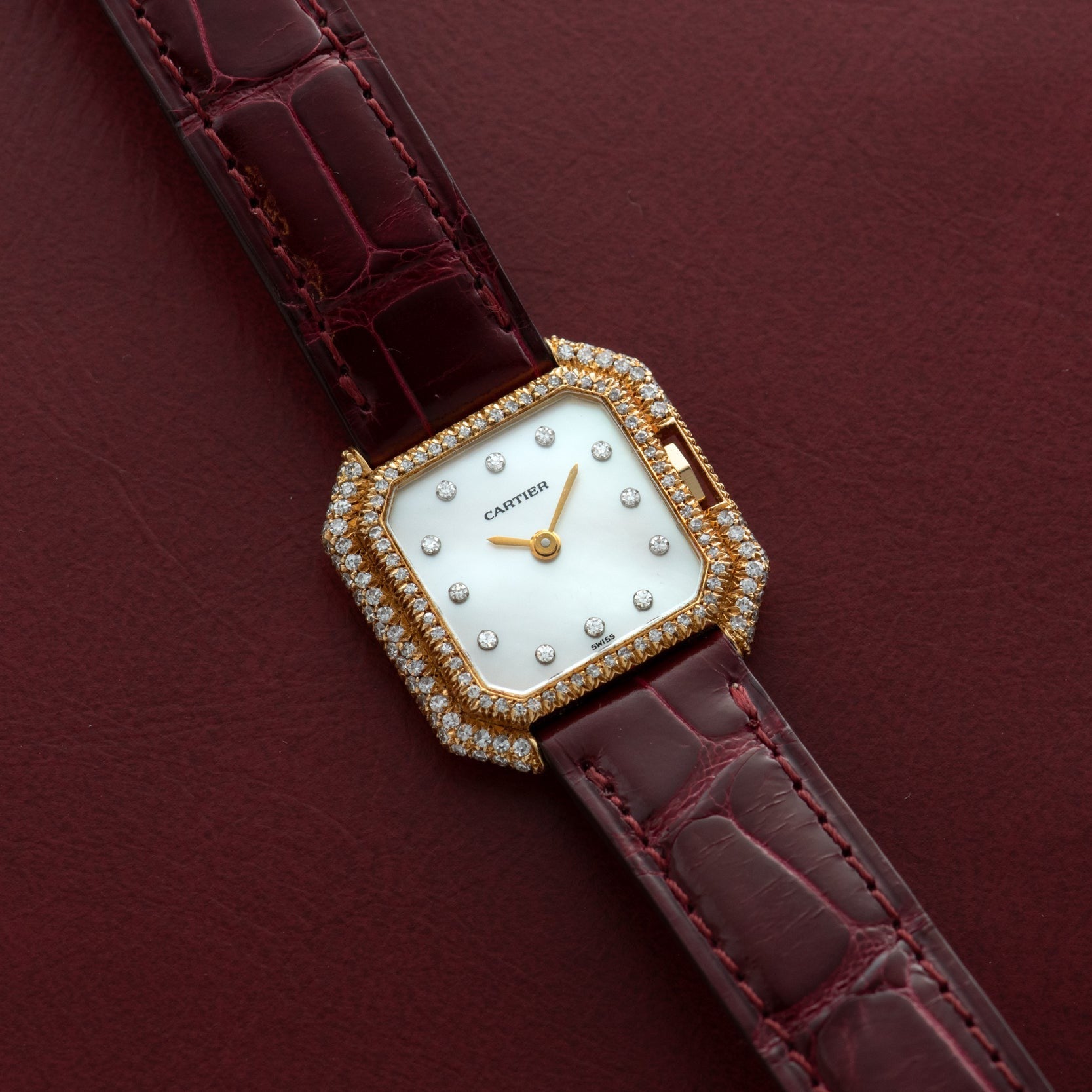The Cartier Ceinture Diamond watch features diamond accents and a burgundy leather strap, showcased on a matching burgundy surface.