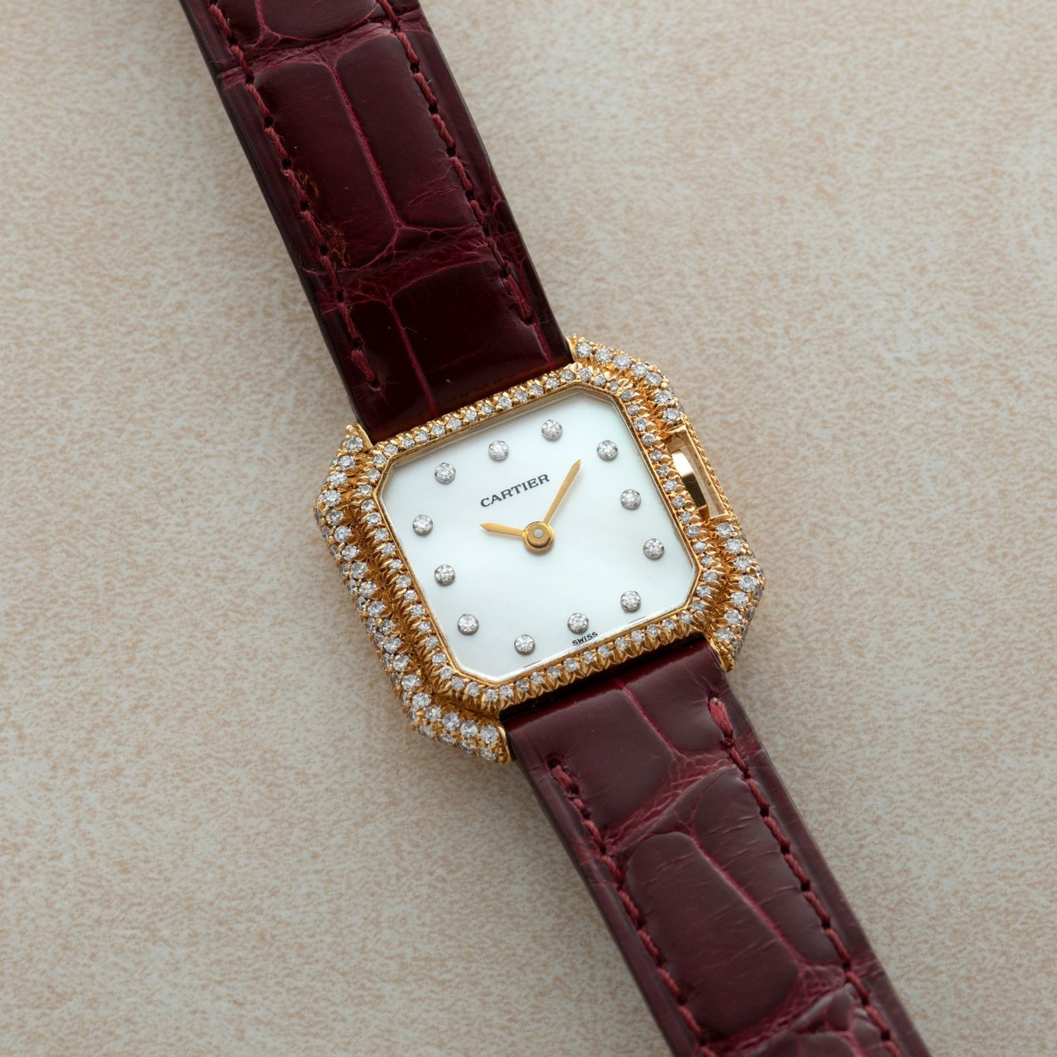 Cartier Ceinture Diamond watch featuring a diamond bezel, white dial, and maroon leather strap displayed on a light surface.