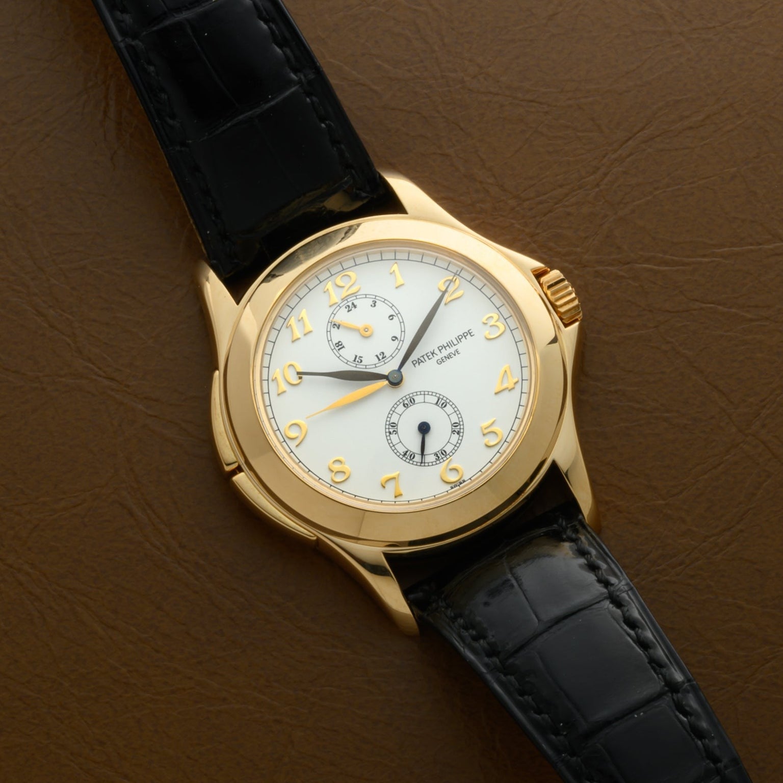 The Patek Philippe Calatrava Travel Time wristwatch with a black leather strap rests on a brown surface, featuring Arabic numerals and subdials.