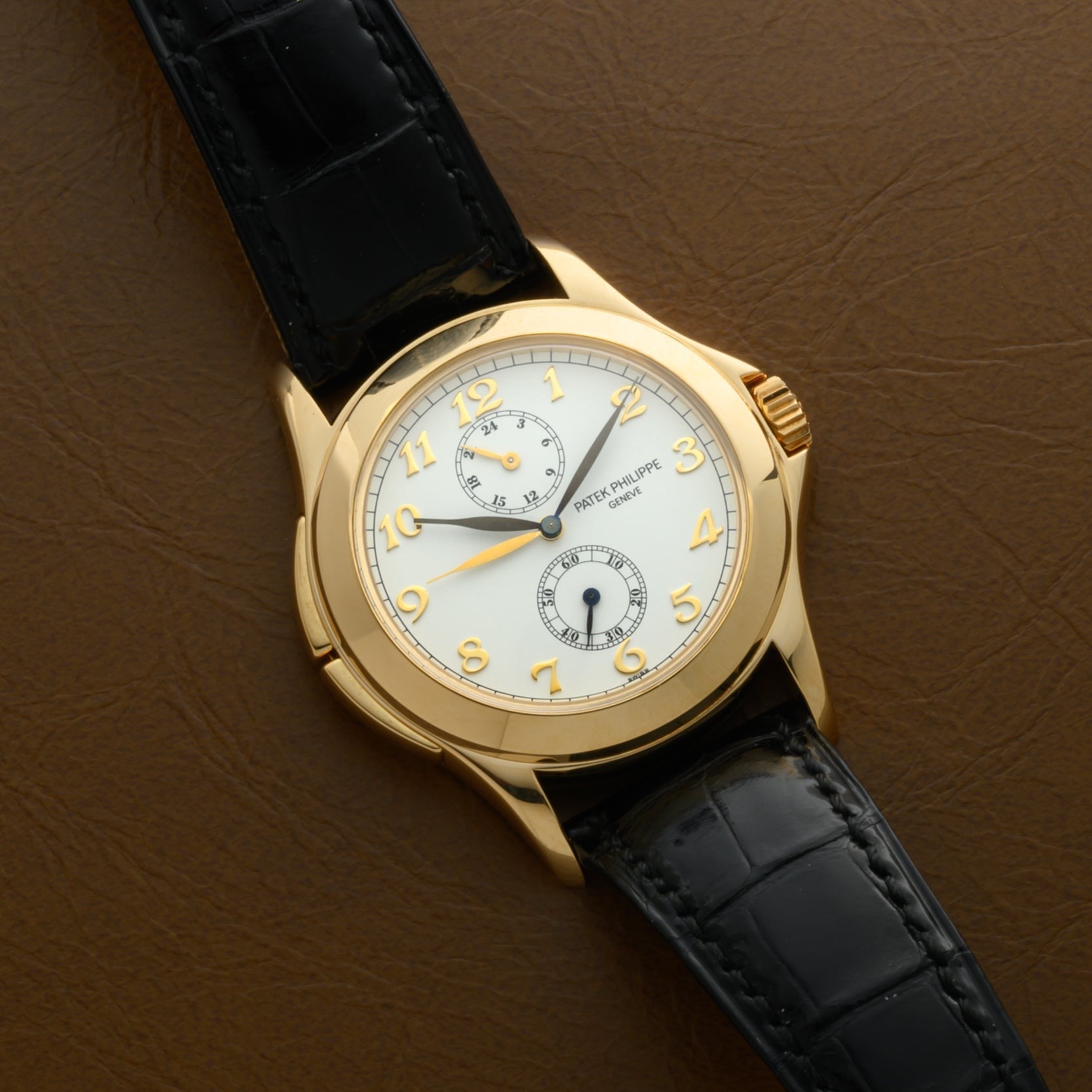 The Patek Philippe Calatrava Travel Time wristwatch with a black leather strap rests on a brown surface, featuring Arabic numerals and subdials.