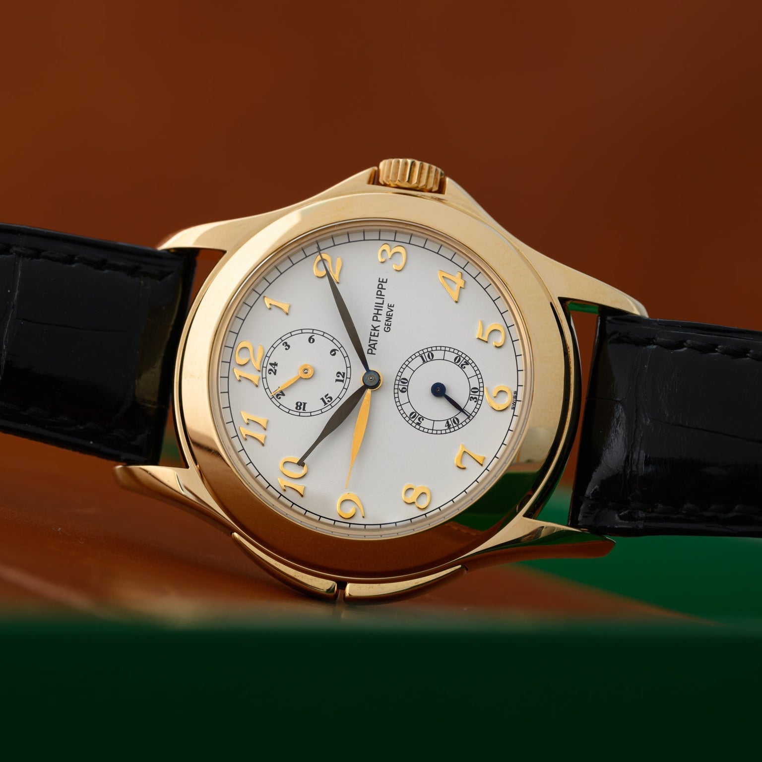 The Patek Philippe Calatrava Travel Time features a gold case, white dial, black leather strap, and Arabic numerals, set against a brown background.