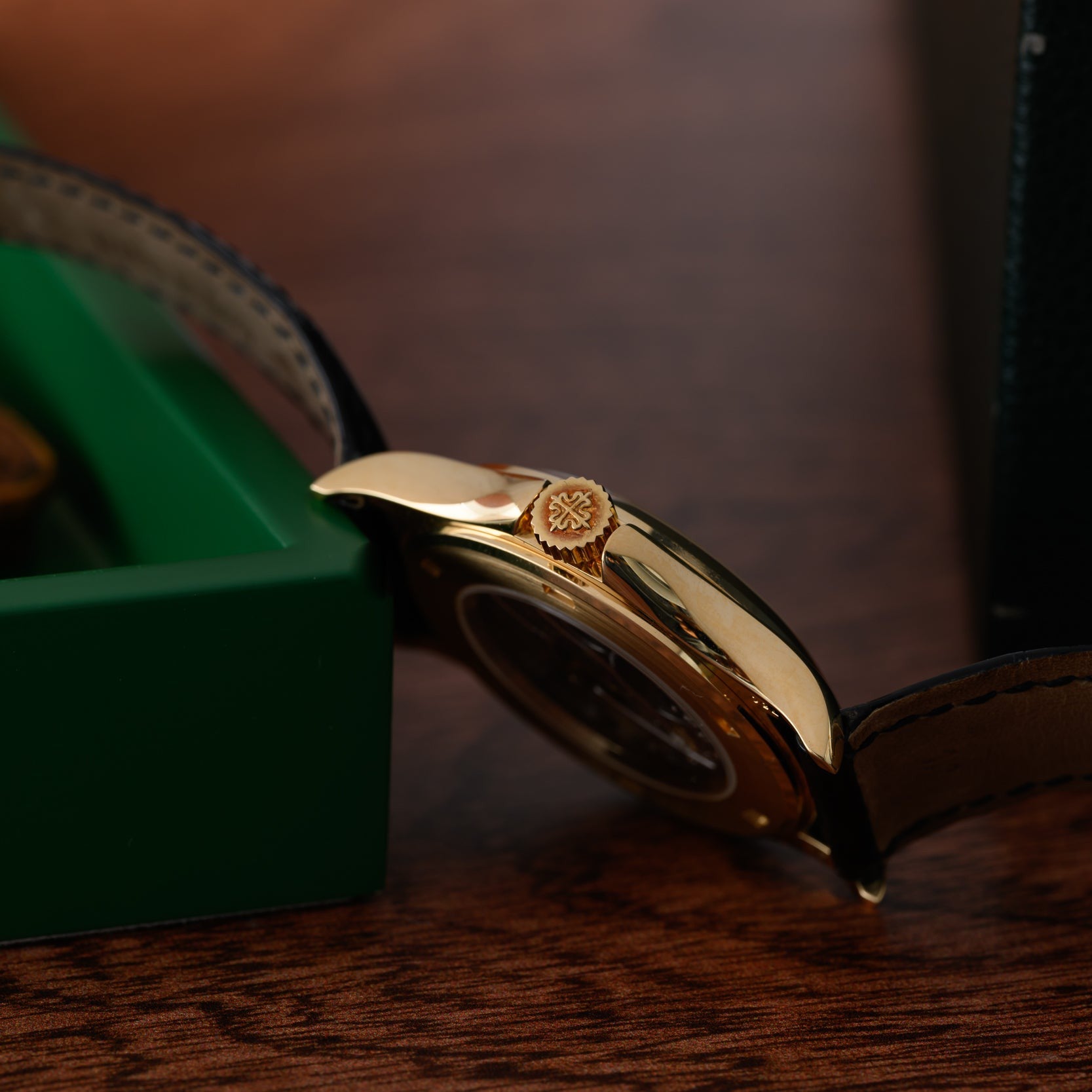 The Patek Philippe Calatrava Travel Time gold wristwatch with a black leather strap rests on a wooden surface beside a green box.
