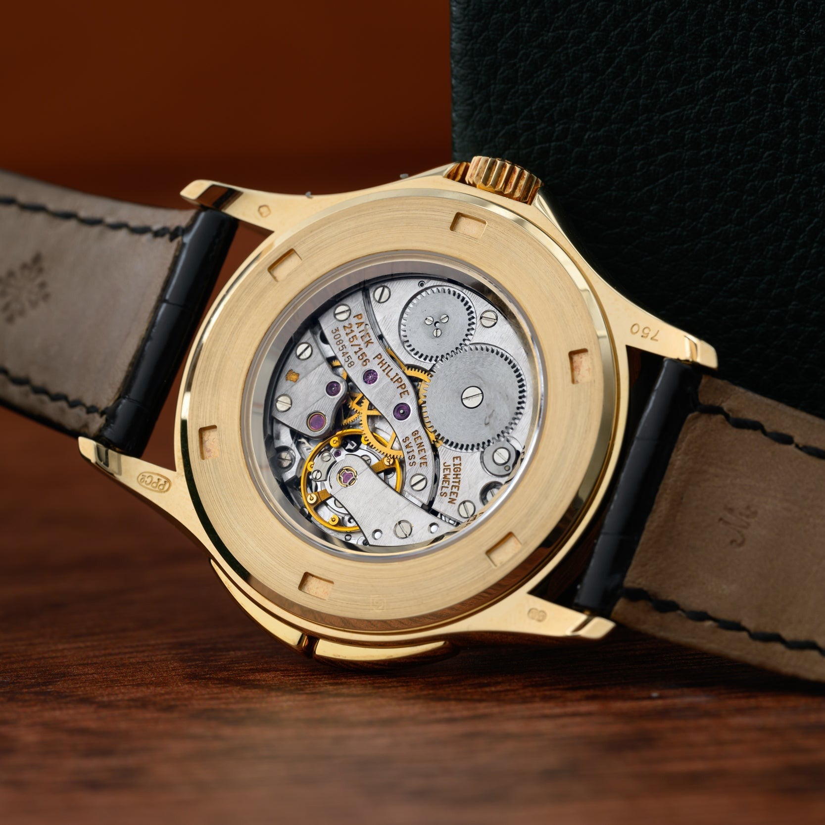 The Patek Philippe Calatrava Travel Time features an exposed mechanical movement, black leather strap, and a green textured background.