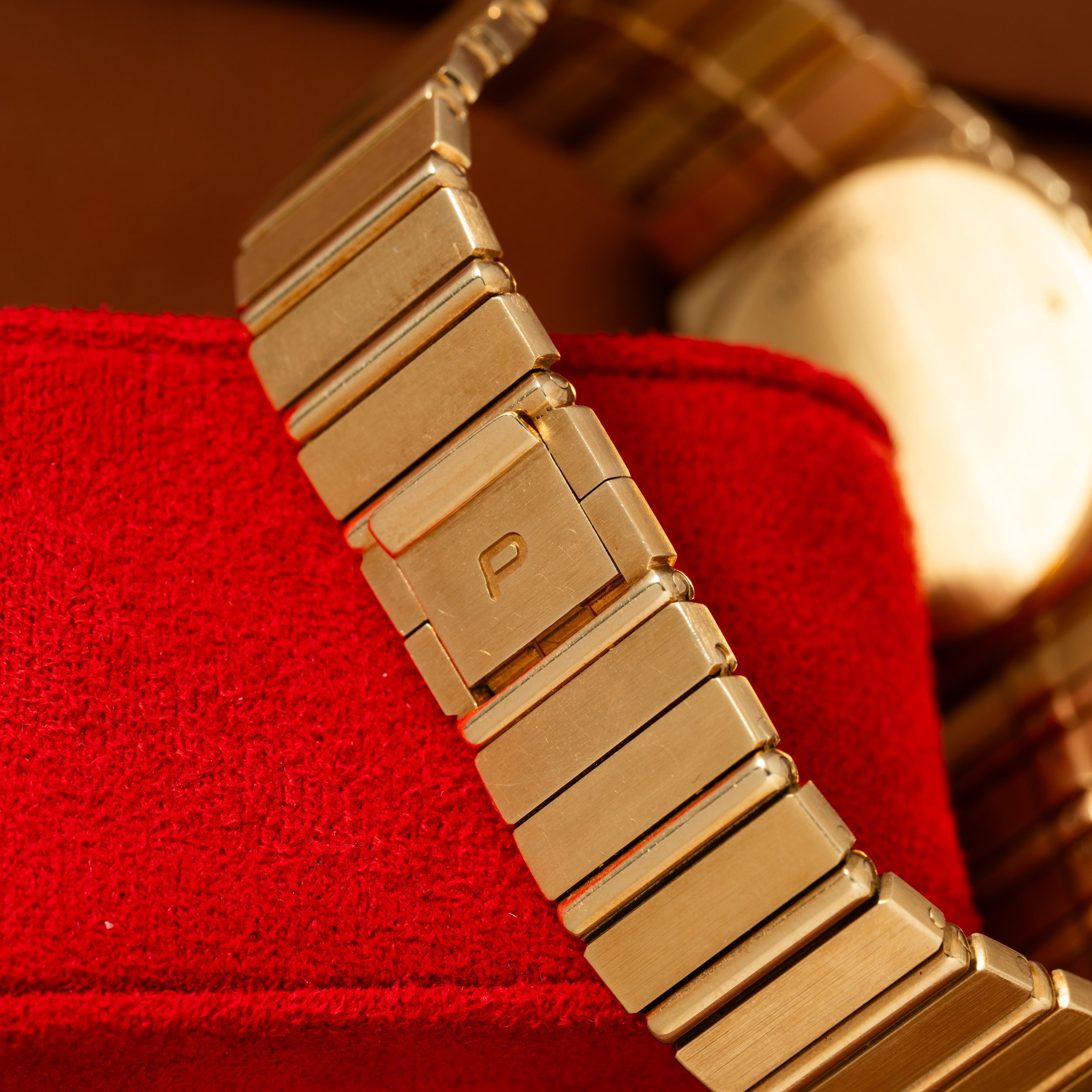 Close-up of a Piaget Polo Calendar gold metal watch band with a P engraved on the clasp, resting on red fabric.