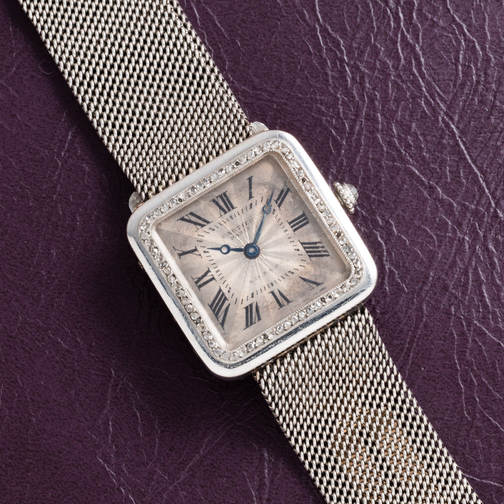 A Cartier Santos Platinum square silver watch with a mesh band and Roman numerals, displayed on a textured dark purple surface.