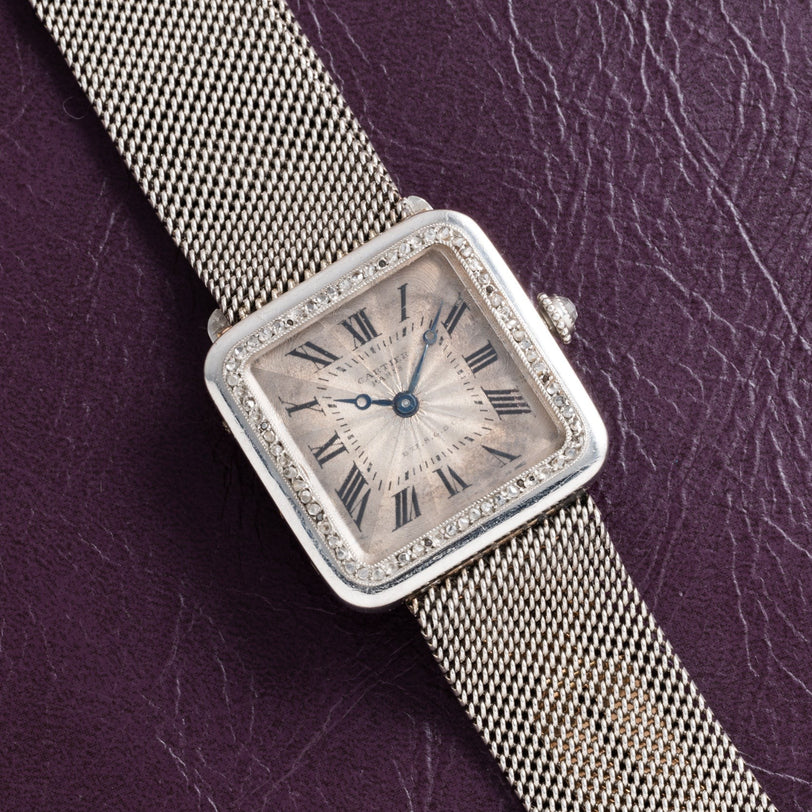 A Cartier Santos Platinum square silver watch with a mesh band and Roman numerals, displayed on a textured dark purple surface.