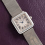 A Cartier Santos Platinum square silver watch with a mesh band and Roman numerals, displayed on a textured dark purple surface.