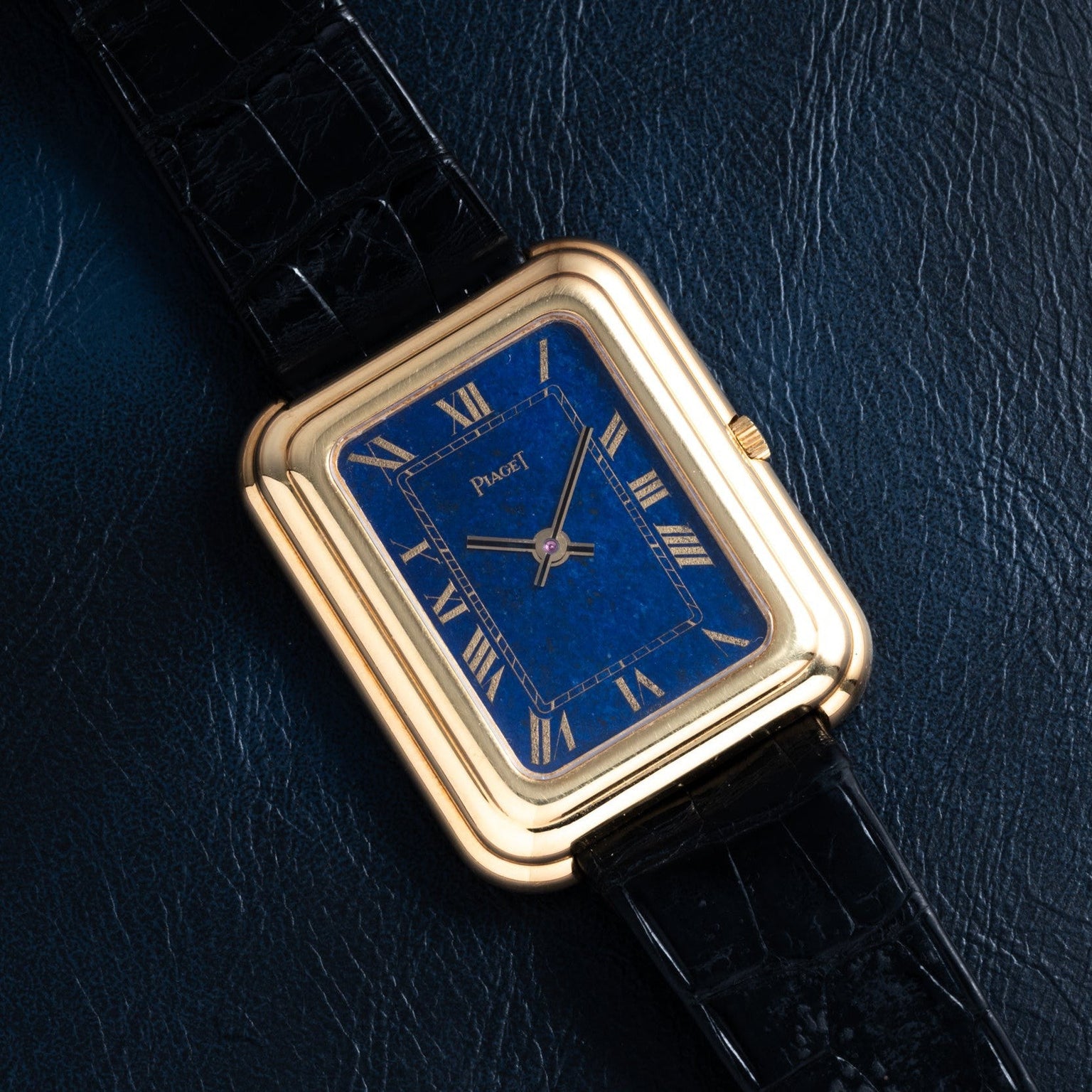 The Piaget Tank Blue Lapis features a gold case, blue rectangular dial with Roman numerals, and a black leather strap, shown on dark leather.