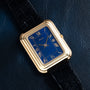 The Piaget Tank Blue Lapis features a gold case, blue rectangular dial with Roman numerals, and a black leather strap, shown on dark leather.