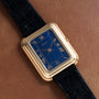 The Piaget Tank Blue Lapis watch features a gold rectangular case, blue face, and black leather strap, displayed on a brown leather surface.