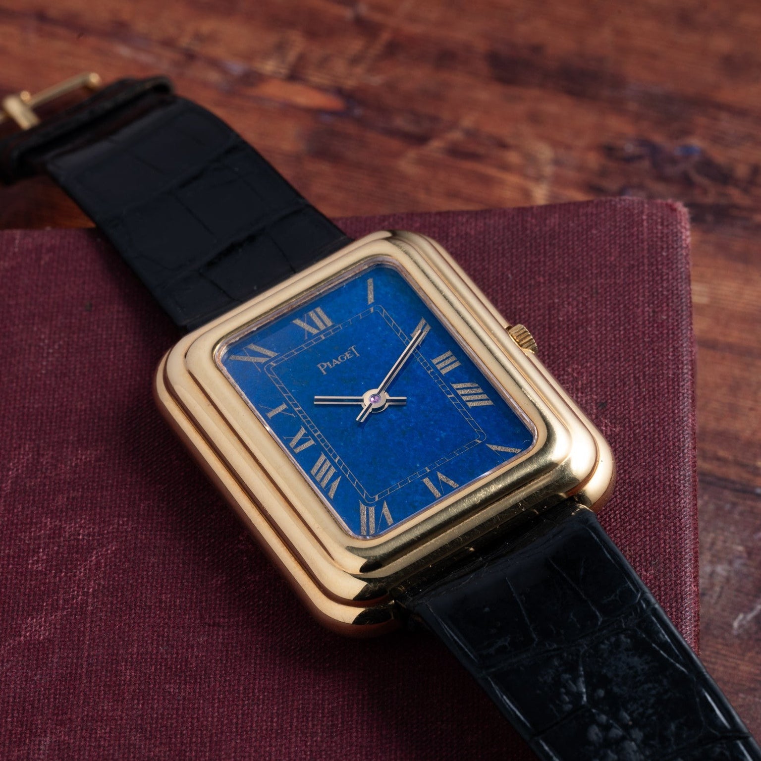 A Piaget Tank Blue Lapis wristwatch with a blue lapis dial and black leather strap rests on a maroon book.