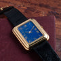 A Piaget Tank Blue Lapis wristwatch with a blue lapis dial and black leather strap rests on a maroon book.