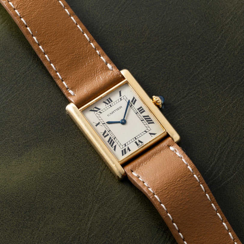 A Cartier Tank Louis gold watch with a brown leather strap rests on a dark green surface.