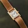 A Cartier Tank Louis gold watch with a brown leather strap rests on a dark green surface.