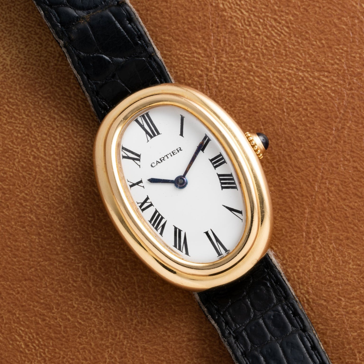 Cartier Baignoire gold wristwatch with an oval face, Roman numerals, and a black leather strap displayed on a brown surface.