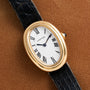 Cartier Baignoire gold wristwatch with an oval face, Roman numerals, and a black leather strap displayed on a brown surface.