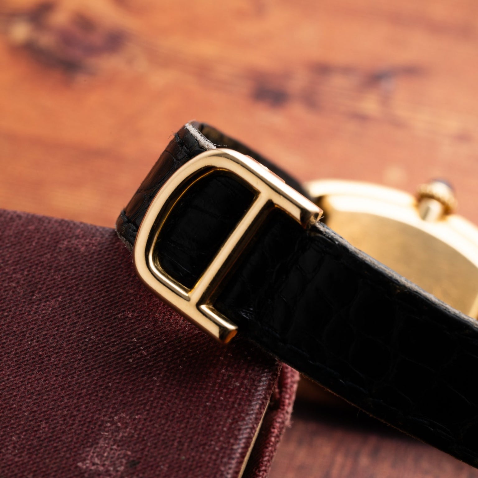 Close-up of the Cartier Baignoire black leather watch strap with a gold buckle, displayed on a maroon textured surface.
