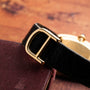 Close-up of the Cartier Baignoire black leather watch strap with a gold buckle, displayed on a maroon textured surface.
