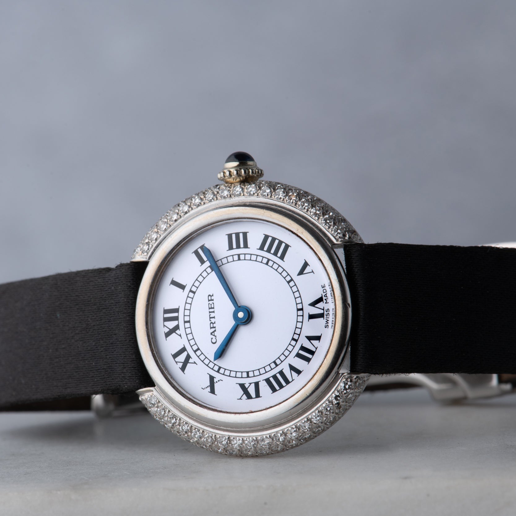 A Cartier Vendome Diamond watch with Roman numerals, a diamond bezel, and a black strap is shown on a gray background.