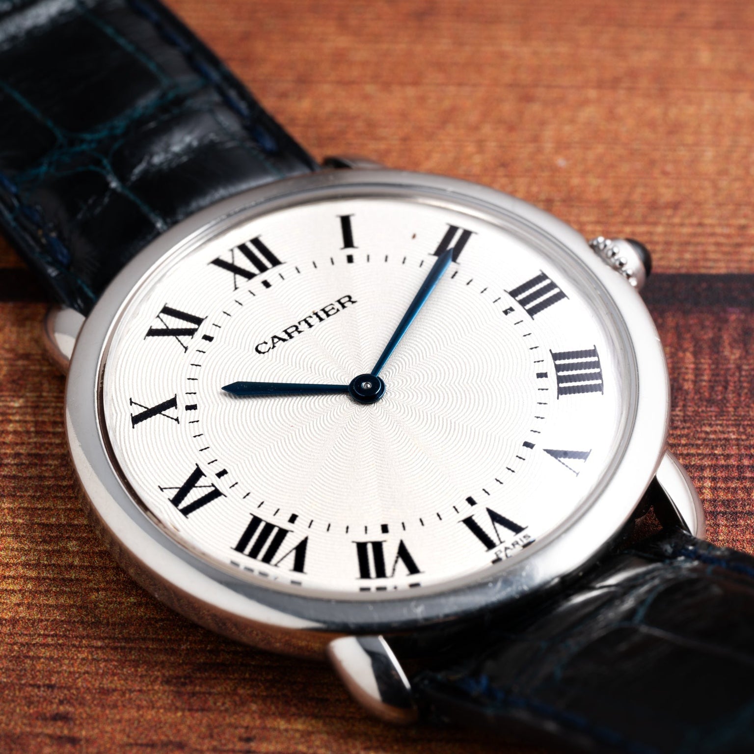 A Cartier Vendome Ronde Louis wristwatch with a white dial, Roman numerals, and blue hands on a leather strap, displayed on wood.