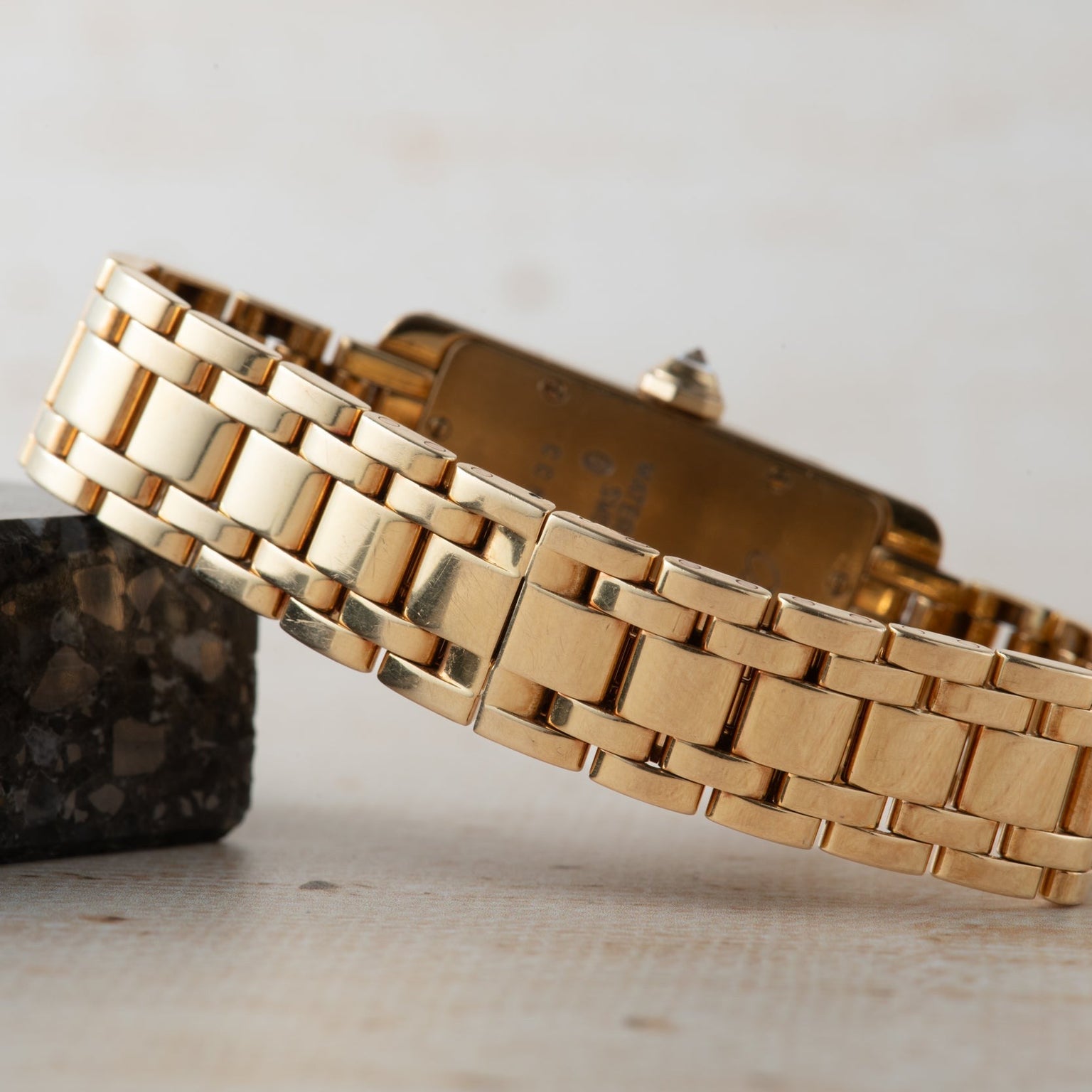 The Cartier Tank Americaine Diamond gold watch with a rectangular face rests sideways, its band displayed, against a small dark stone.