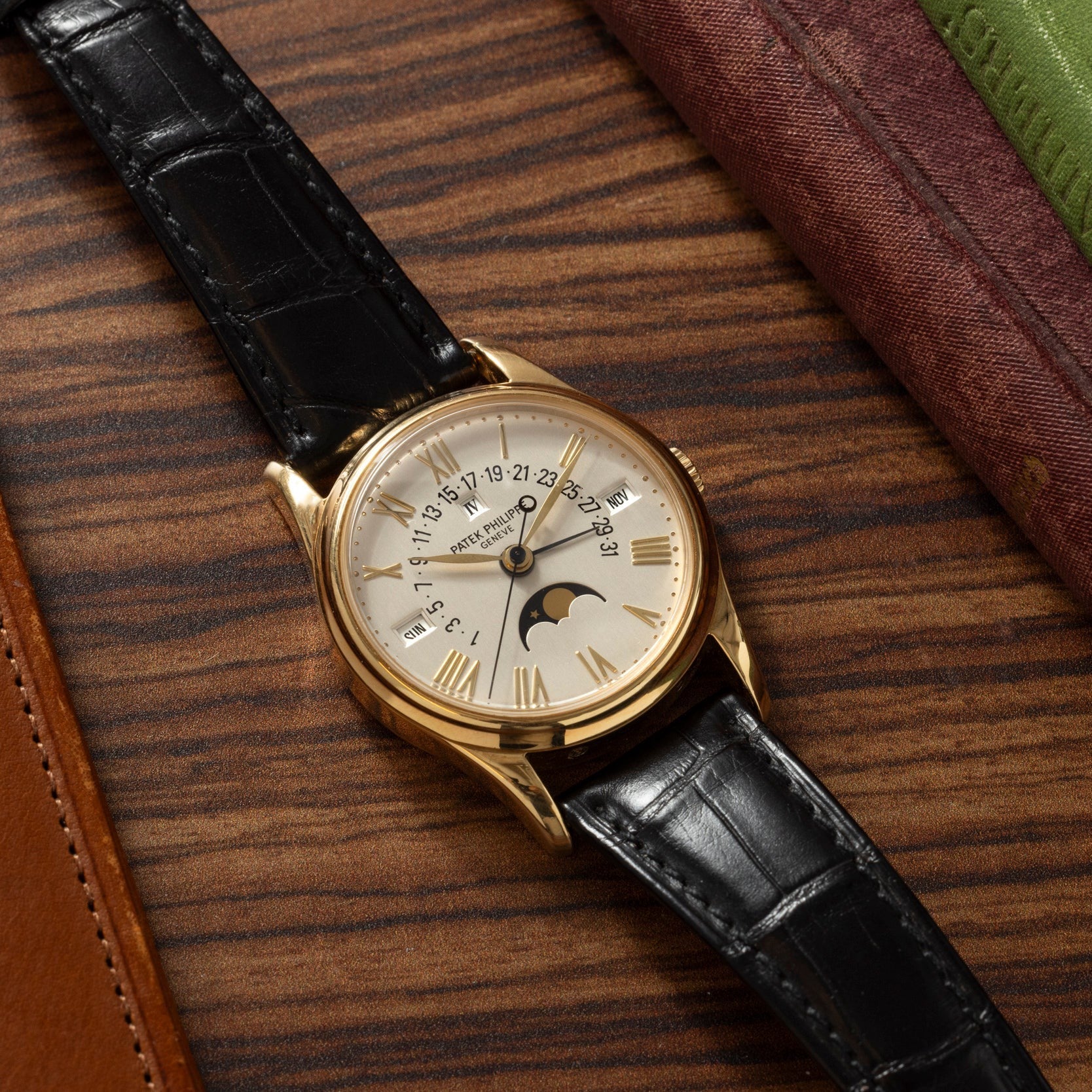 The Patek Philippe Retrograde Perpetual Calendar wristwatch, featuring a black leather strap, is displayed on a wooden surface beside brown and green books.