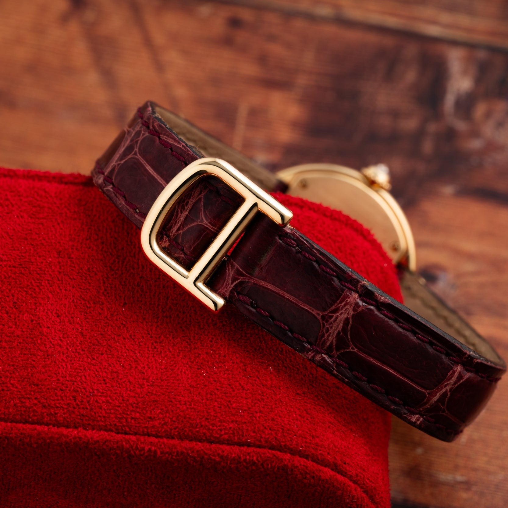 The Cartier Ellipse, a brown leather watch with a gold buckle, rests on a red suede surface against a wooden background.