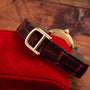 The Cartier Ellipse, a brown leather watch with a gold buckle, rests on a red suede surface against a wooden background.