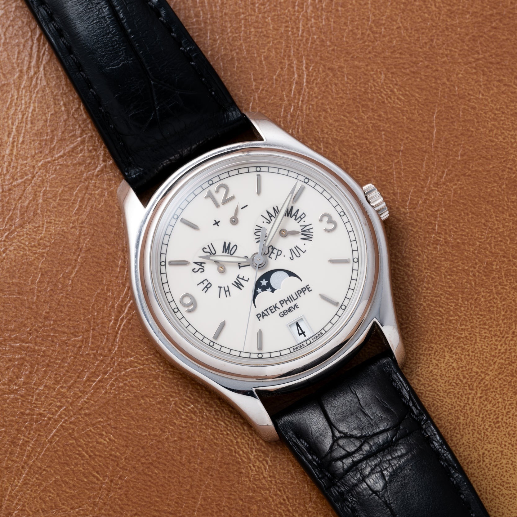 The Patek Philippe Annual Calendar Moonphase wristwatch features a black leather strap, elegantly displayed on a brown leather surface.
