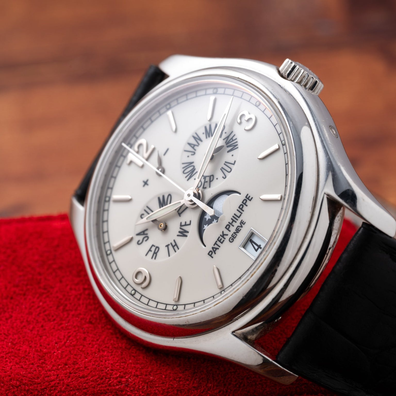 A Patek Philippe Annual Calendar Moonphase wristwatch with a silver case and black leather strap rests on a red cloth, displaying the date and month.