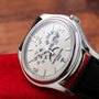 A Patek Philippe Annual Calendar Moonphase wristwatch with a silver case and black leather strap rests on a red cloth, displaying the date and month.