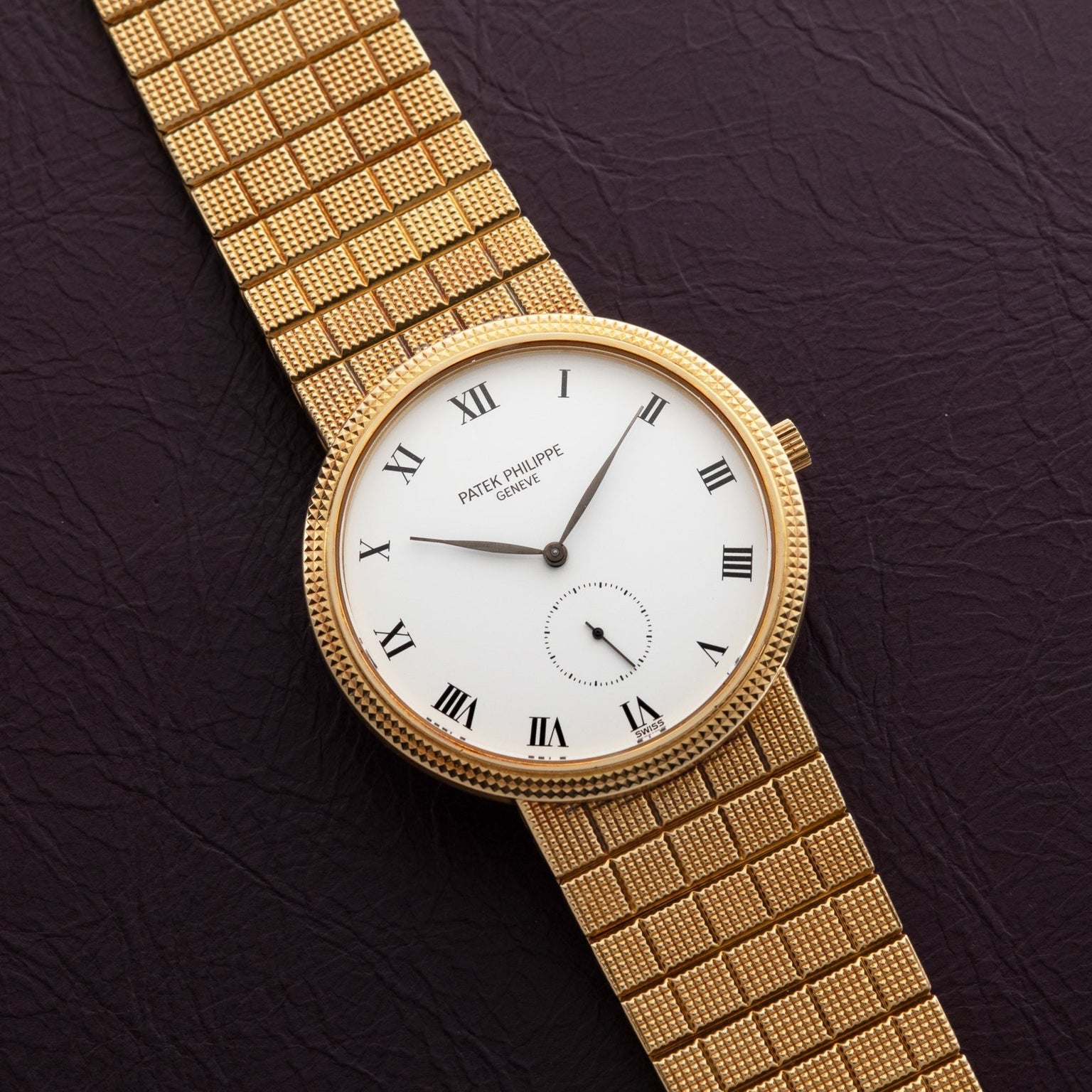 Patek Philippe Calatrava gold watch with a white face, Roman numerals, and mesh band displayed on a dark textured surface.