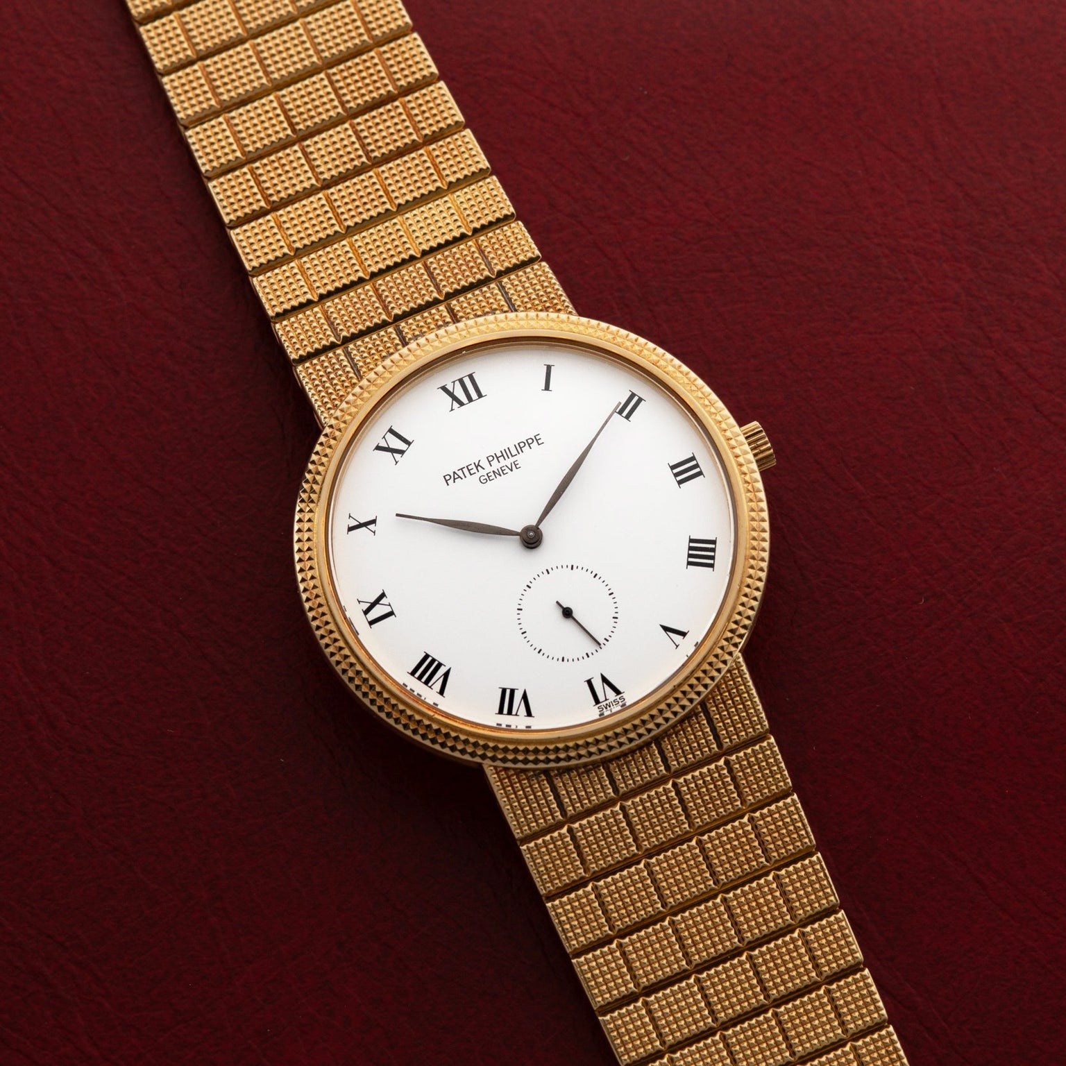 The Patek Philippe Calatrava gold wristwatch features a white Roman numeral dial and a textured band, displayed on a red leather surface.