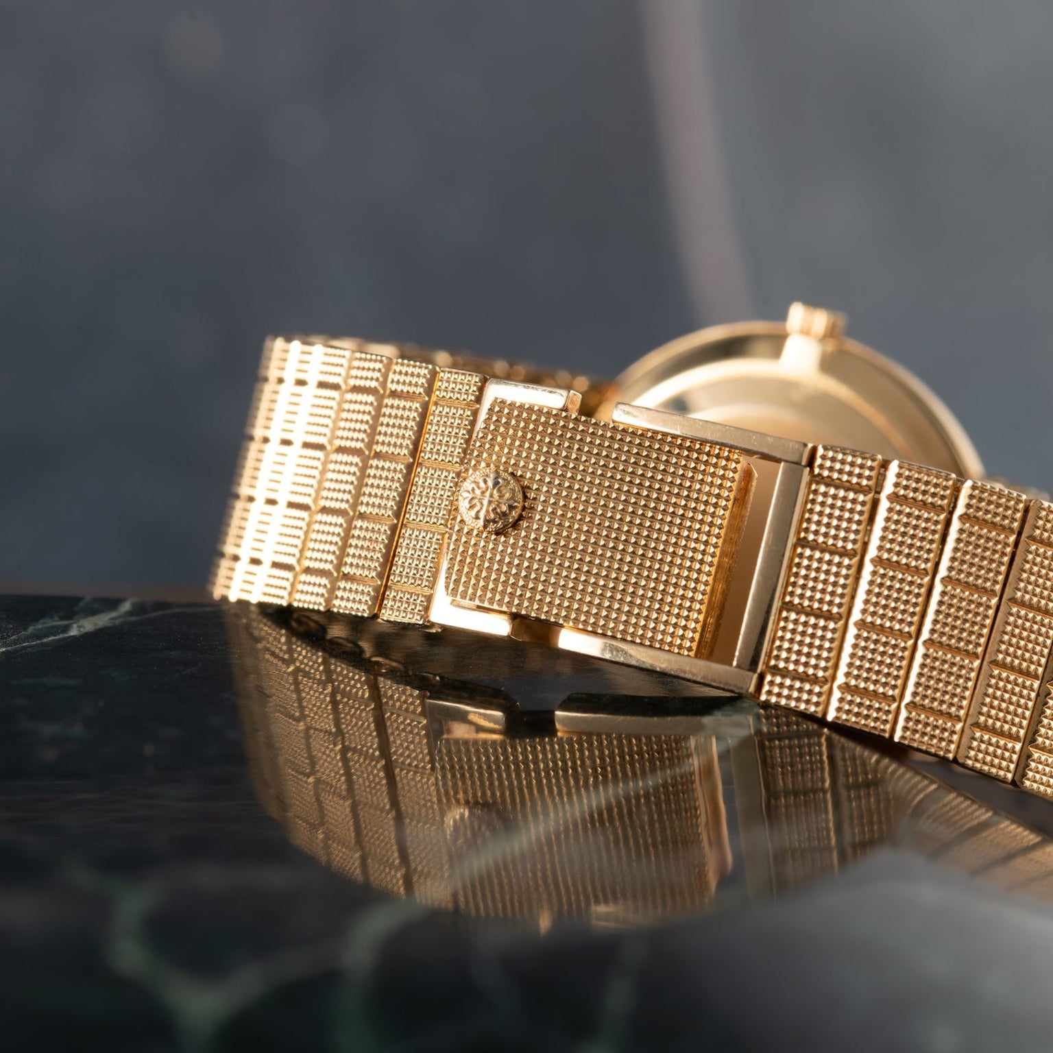 Close-up of the Patek Philippe Calatrava gold watch with a textured metal band, resting on a glossy dark marble surface.