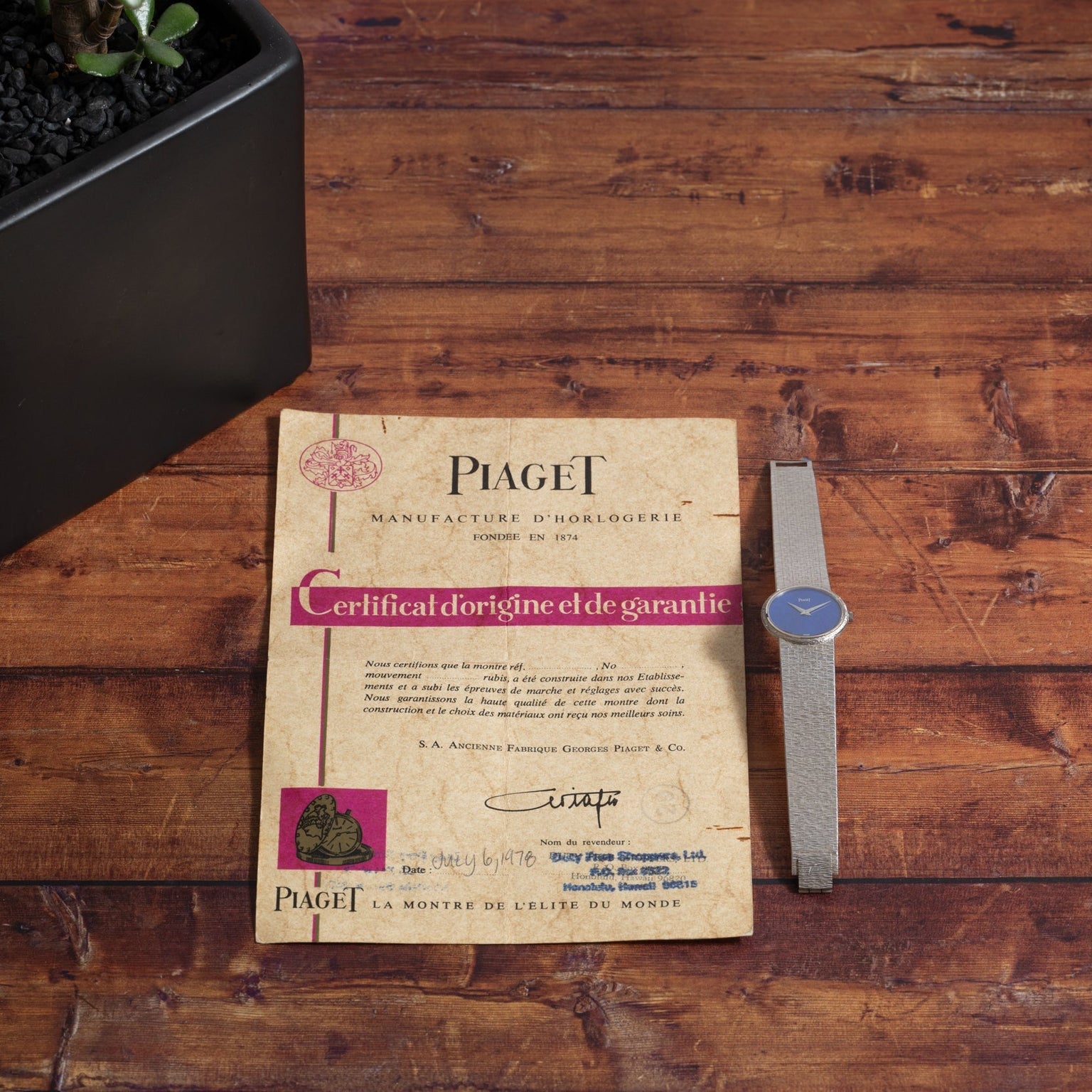 A Piaget White Gold Lapis Lazuli Dress Watch and certificate rest on a wooden table next to a black plant pot.